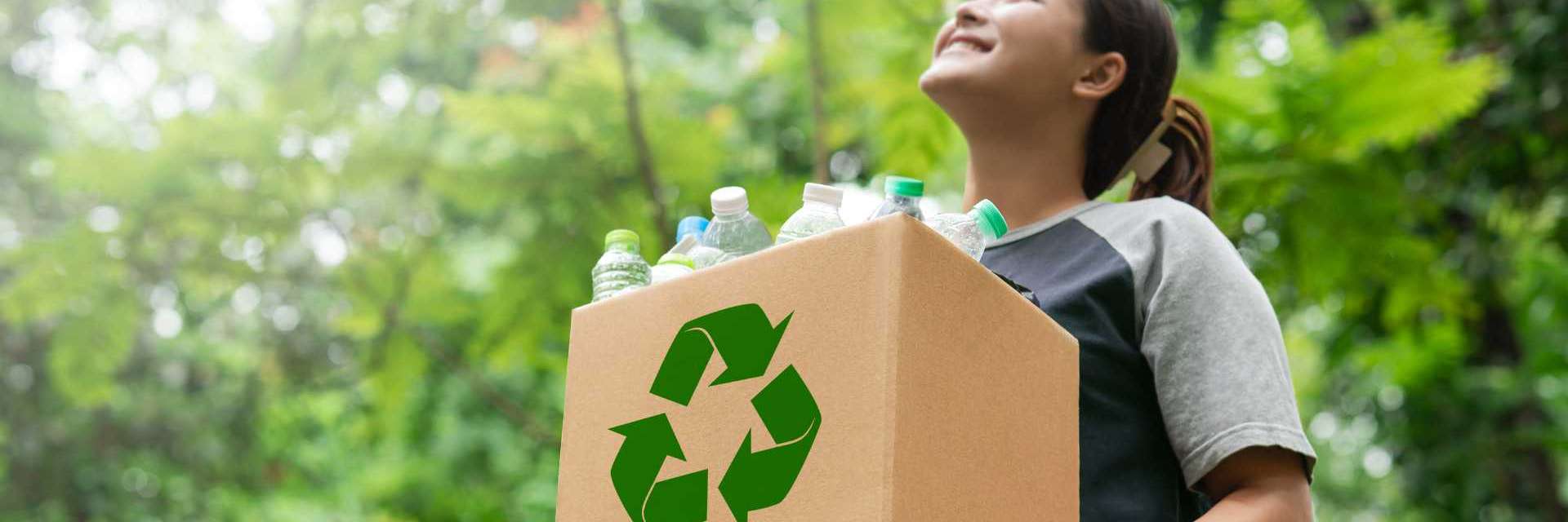 pessoa segurando uma caixa de papelão com o símbolo de reciclagem, cheia de garrafas plásticas, em um ambiente natural com árvores ao fundo.