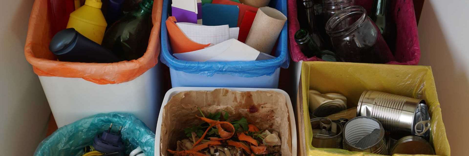 A imagem mostra um conjunto de cinco lixeiras coloridas para a separação de recicláveis: plásticos em uma lixeira com saco laranja, papéis em azul, vidros em rosa, compostos orgânicos em um recipiente branco e metais em um saco amarelo.